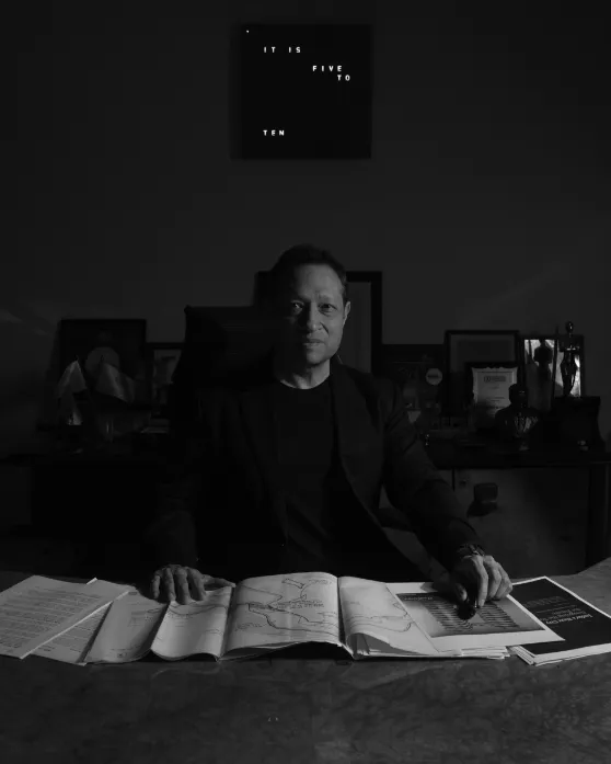 Jimmy Mistry at his desk with township plans and awards behind him, black-and-white portrait